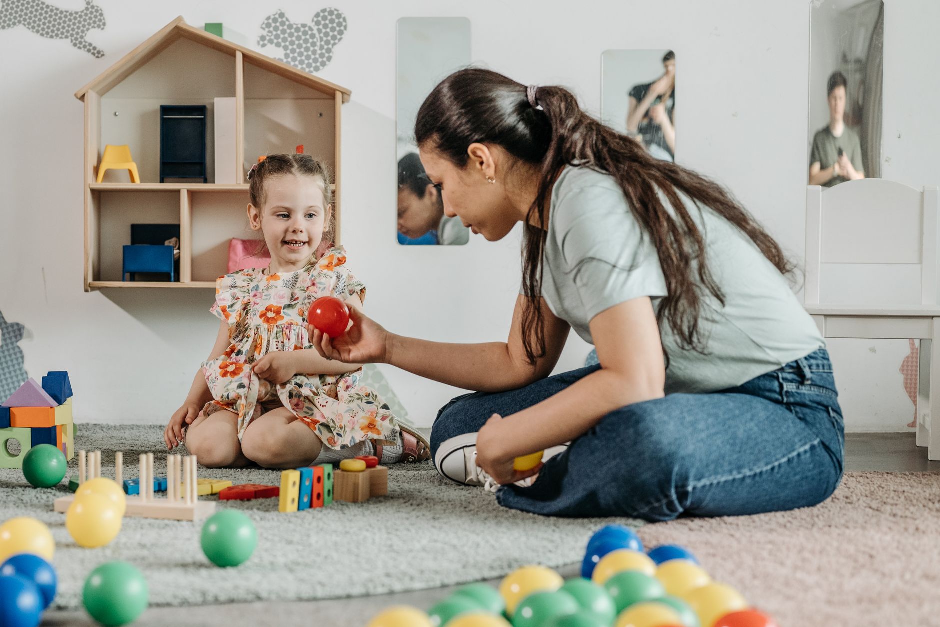 A teacher sitting on the floor playing quietly with a young child in a sunlit classroom